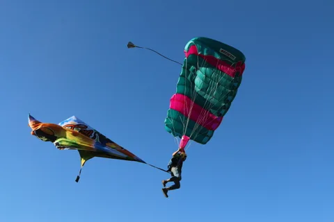 A skydiver trails a colorful banner beneath a teal and pink parachute against deep blue sky.
