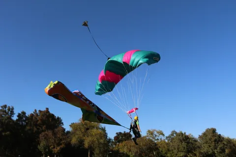 A skydiver with a trailing banner descends toward the treeline with a parachute flared overhead.