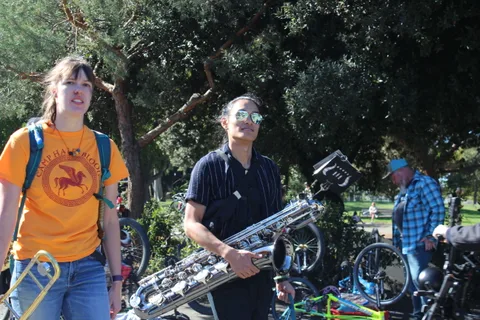 A trombone player performs with a saxophonist beside him under the trees.