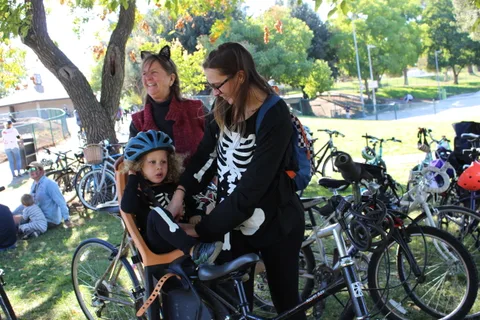 A mother buckles her costumed toddler into a bike seat while a grandmother looks on.