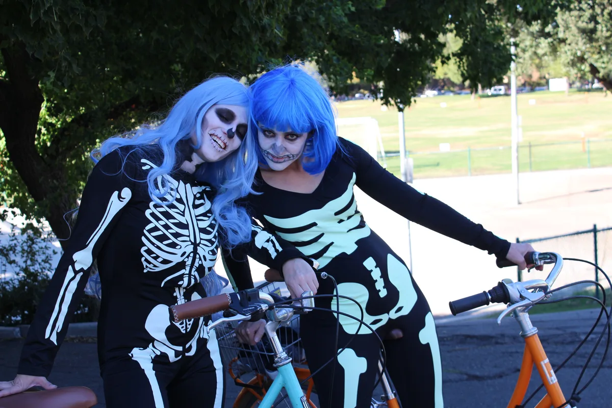 Two riders in blue wigs and skeleton face paint lean together on their bikes.