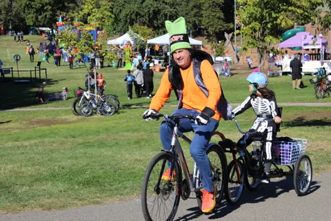 A man in a Goofy hat rides beside a child in a skeleton costume on an adaptive trike.
