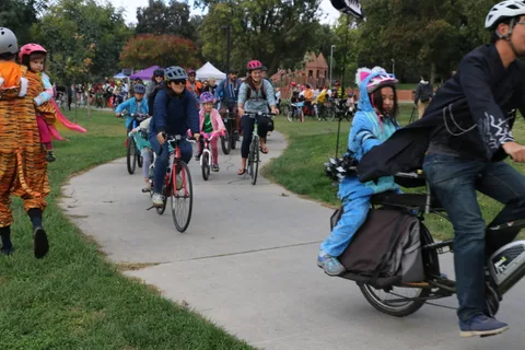 Costumed riders in helmets pedal down a park path at the start of the parade.