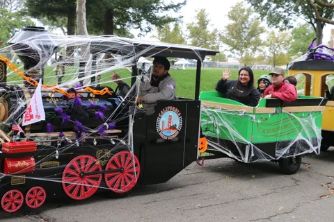 A driver steers a cobweb-covered trackless train as a waving family rides in the car behind him.