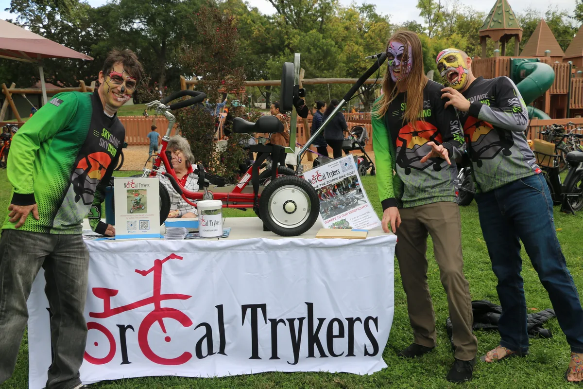 Three face-painted volunteers stand behind the NorCal Trykers donation table with a custom trike on display.