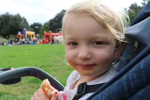 A toddler with food on her cheeks looks toward the camera from a stroller.