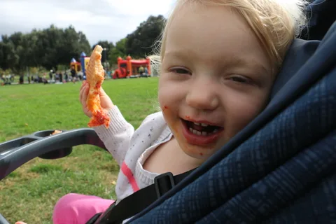 A toddler with a sauce-smeared face grins a gap-toothed smile while holding up a slice of pizza.