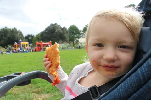 A toddler with a messy face holds up a snack in a park setting.