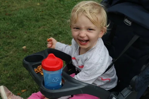 A toddler grins with a sippy cup in her stroller tray.