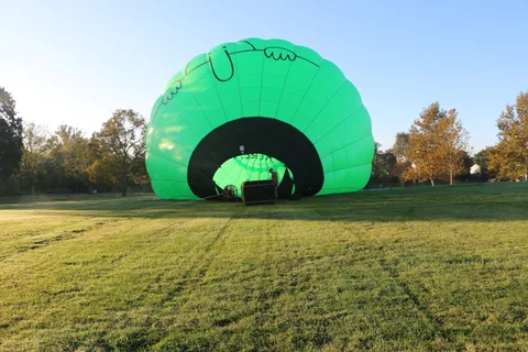 A fully inflated green hot air balloon with a whimsical painted face glows in a grassy field at sunrise.