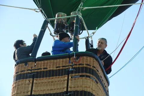 Riders in the balloon basket smile and reach toward the camera mid-flight.