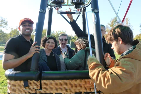 A group of guests smile together near the hot air balloon in warm morning light.