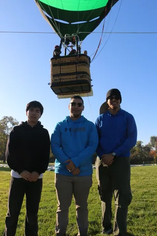 Three young men stand together under the hot air balloon basket.