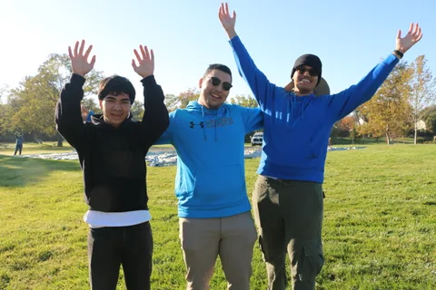 Three friends cheer with arms raised on the grass as the hot air balloon is laid out behind them.