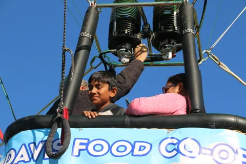 A boy rides in the hot air balloon basket beside the Davis Food Co-op banner, reaching toward the controls.