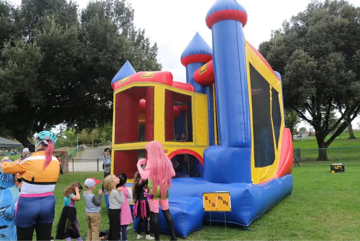 A tall red, blue and yellow castle bounce house on the grass with a queue of children waiting.
