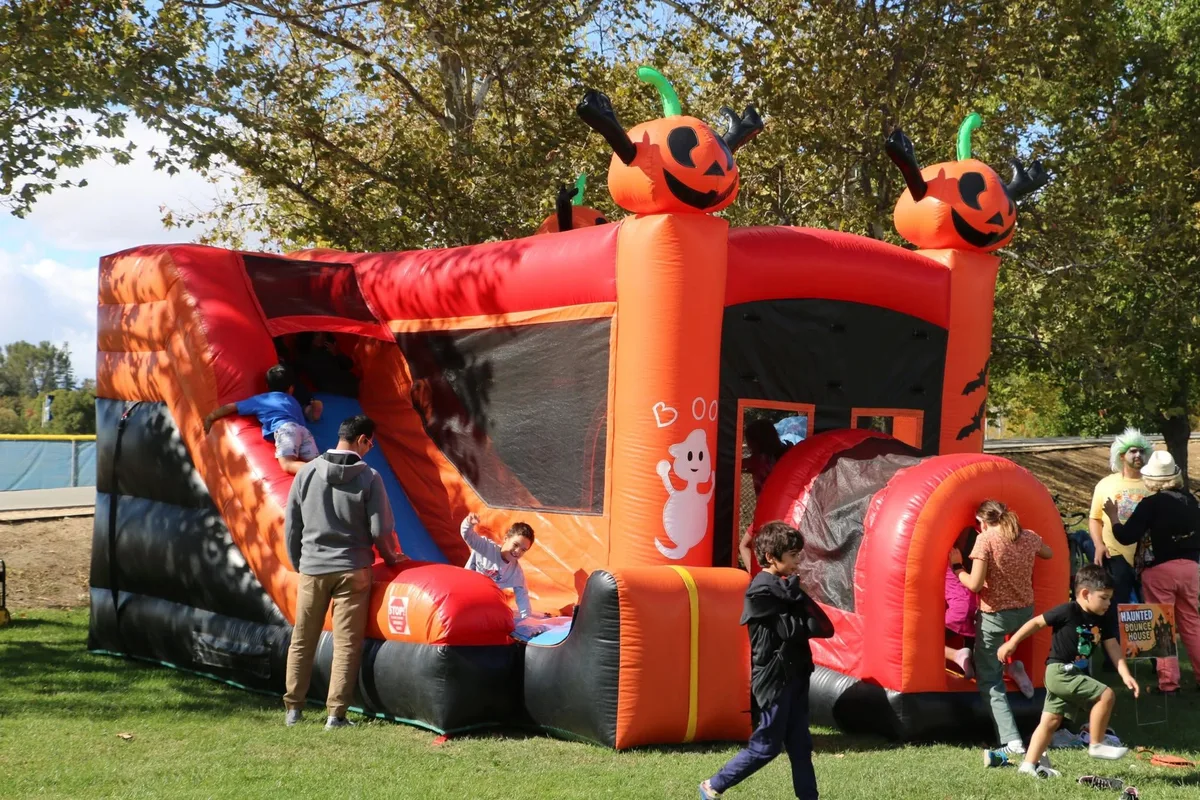 Children climb and slide on a large orange Halloween bounce house topped with grinning pumpkins.