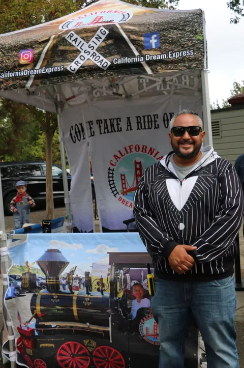 Surf, the founder and owner of California Dream Express, standing in front of his Come Take a Ride On banner beside a painted train panel, with a small child watching in the background.