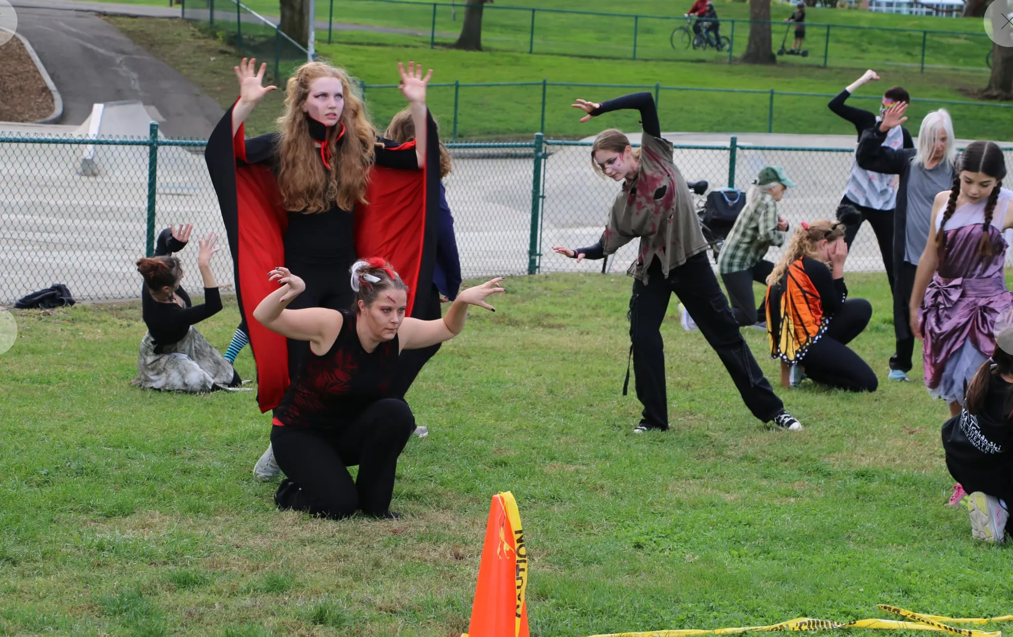 Costumed dancers perform on the grass at Community Park, a caped vampire and a kneeling dancer reaching forward while others hold their positions behind.