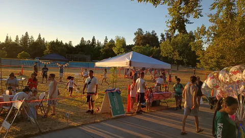Families spread across the Discovery Family Fun Zone in late afternoon light, with a canopy, lawn games, tables and a cluster of balloons.