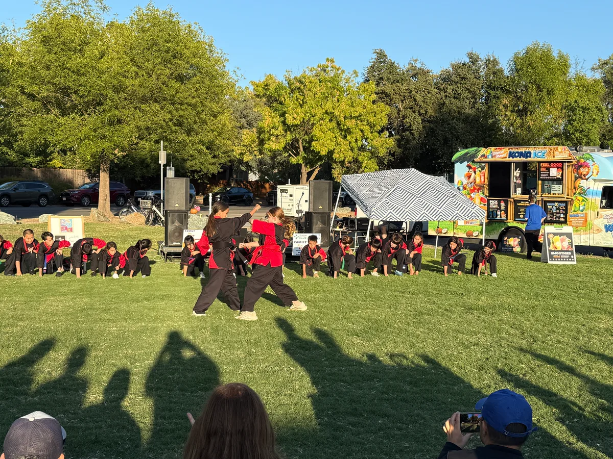 Two young martial artists in black and red uniforms face each other mid-demonstration on a grass field, with a row of teammates kneeling in a line behind them and a Kona Ice truck and canopy at the edge of the field.