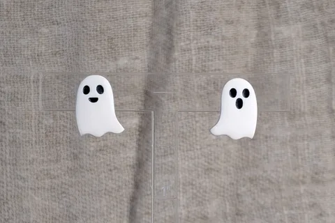 Two small white ghost stud earrings with simple black faces, on a clear display stand against pale linen.