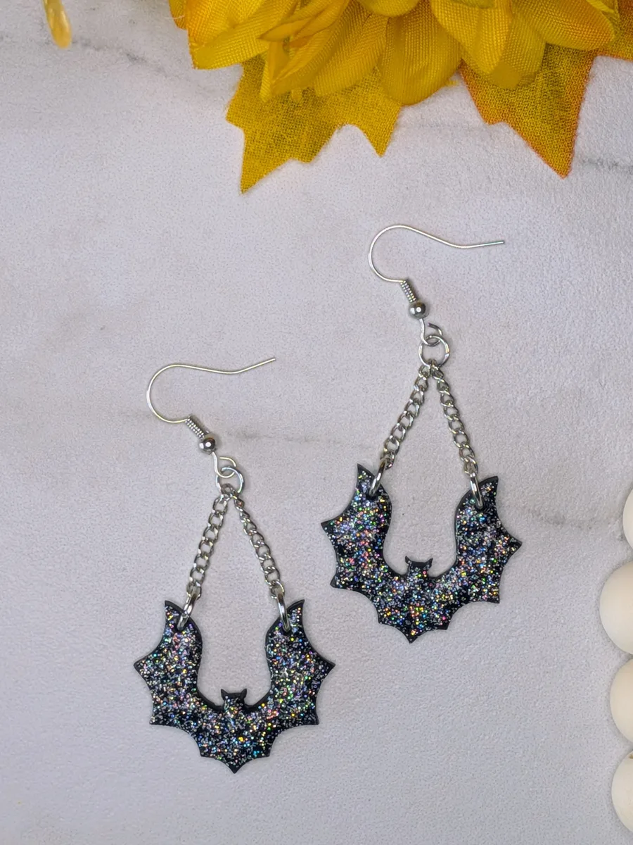 A pair of glittery black bat earrings hanging from silver chains against a pale linen background.