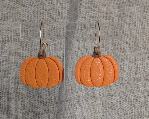 A pair of orange polymer clay pumpkin earrings with fine swirled detail, on a pale linen background.