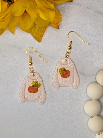Cream coloured polymer clay earrings shaped like knitted sweaters, each with a small orange pumpkin on the front, on a marble surface beside a sunflower.
