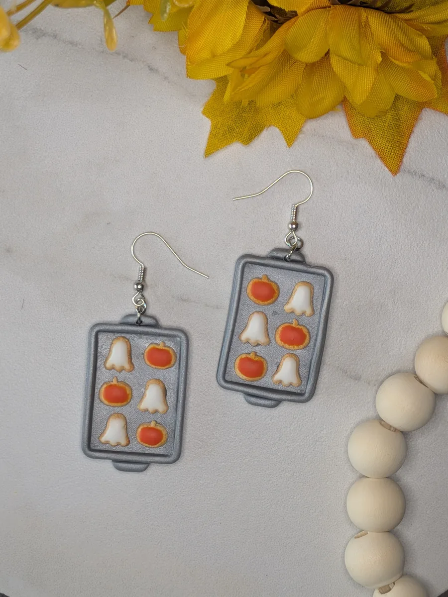 Polymer clay earrings shaped like grey baking trays, each holding tiny white ghost and orange pumpkin cookies, on a marble surface beside a sunflower.
