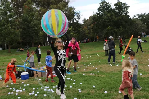 A volunteer in a skeleton suit holds a giant striped beach ball overhead as children play with pool noodles on a field scattered with hundreds of white balls.
