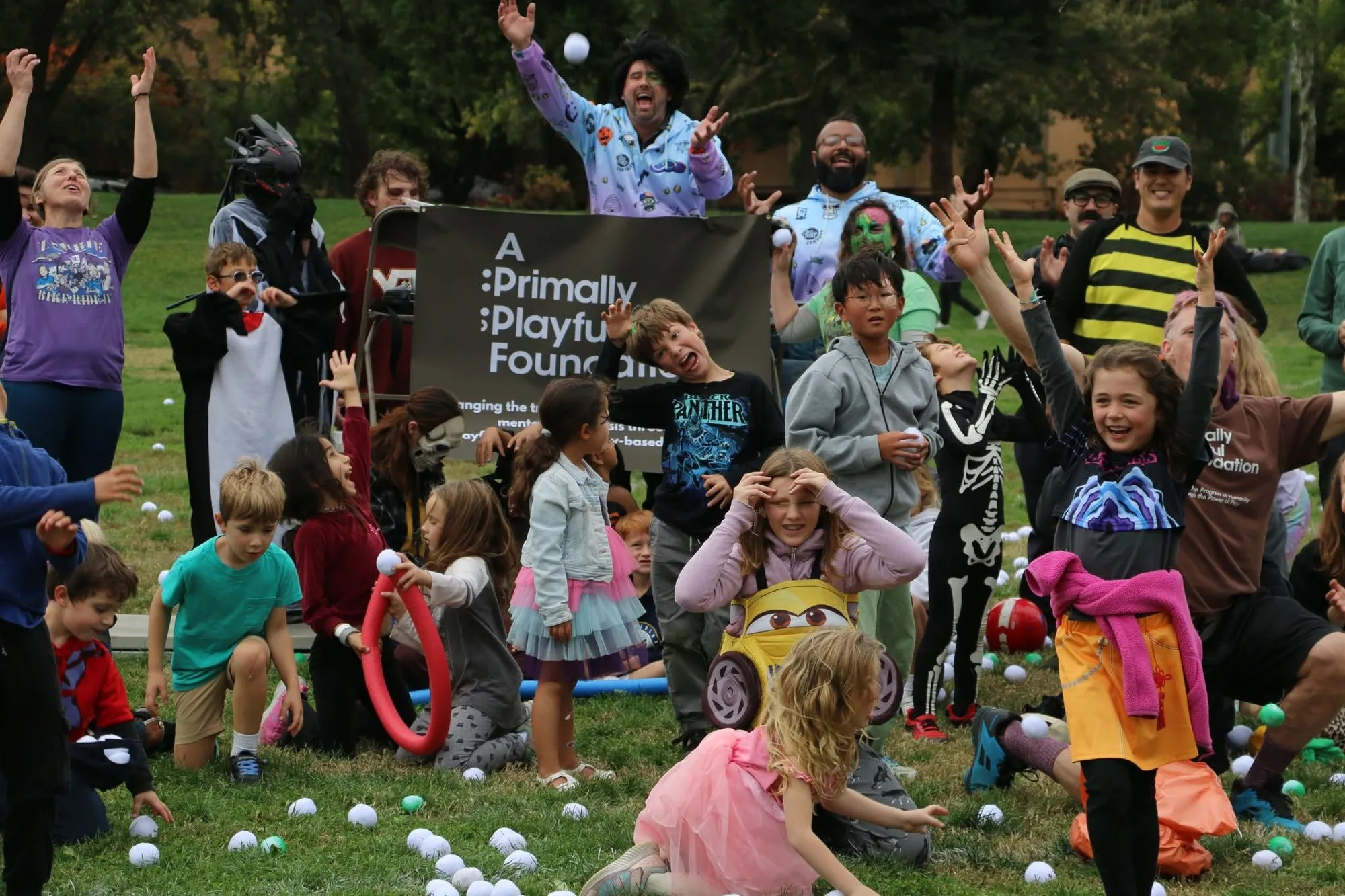 Dozens of children and adults in costume throw their hands in the air in front of a banner reading A Primally Playful Foundation.