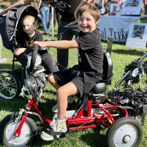 A boy laughs on a red adaptive trike on the grass at the festival, a NorCal Trykers banner behind him.