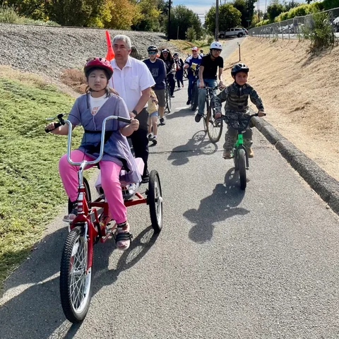 A child on a red adaptive trike rides a paved path as other riders follow behind.