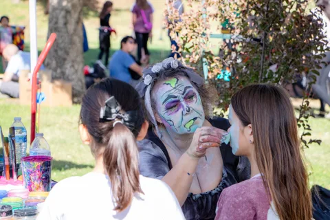 A face painted performer leans in to talk with two visitors at the face painting table.