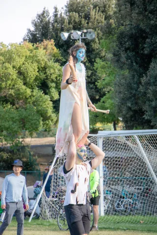 An acrobatic duo hold a standing lift on the grass as families watch.