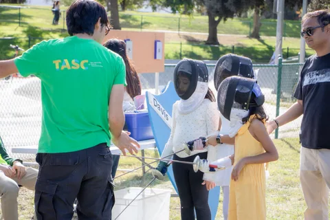 A coach helps two young fencers put on their masks at the fencing booth.