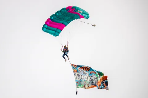 A skydiver steers a bright canopy with a flag trailing behind.