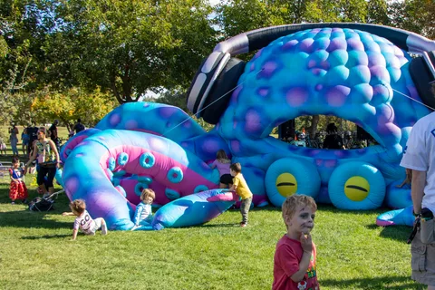 Children play on the grass beside a giant blue and pink octopus bounce house.