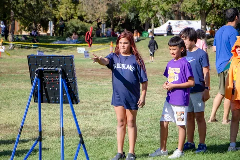 A game attendant stands beside a ball toss frame on the grass as children wait their turn.