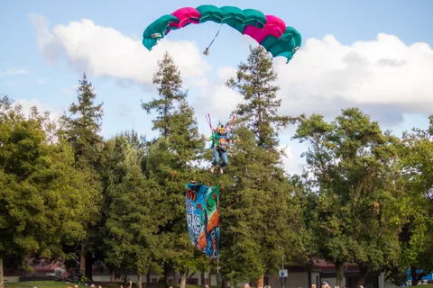 A canopy comes in low over the pines with a flag below it.