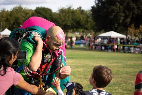 A skydiver in face paint leans down to talk with two children.
