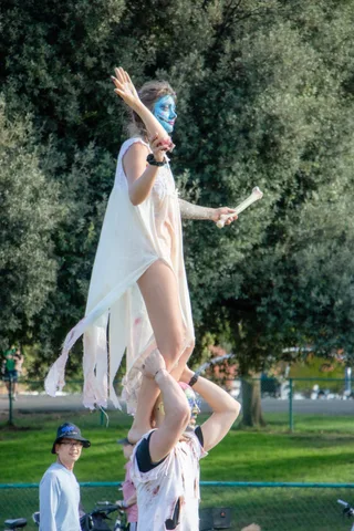 A performer in a flowing gown stands tall on a partner's shoulders under the trees.