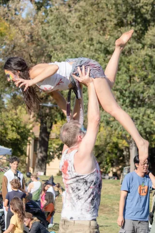 An acrobat is held overhead in a one handed lift while onlookers watch.