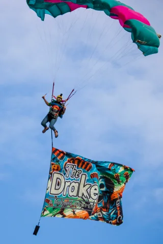A skydiver drops in with a large sponsor flag flying below.
