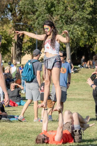 An acrobat stands on a partner who is lying on the grass, arms spread wide.