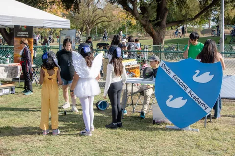 Children in costume try fencing on the grass beside a blue shield banner.