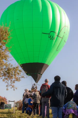 A crowd watches a green balloon inflate on the lawn
