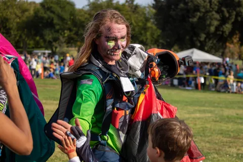 A skydiver in face paint moves through the crowd with gear.