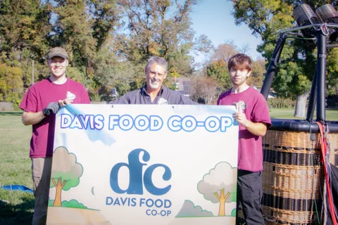 Three people hold a sponsor banner beside a balloon basket